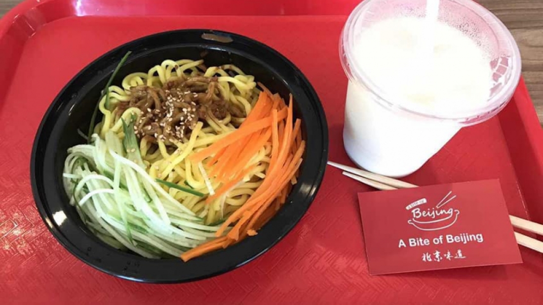 A red cafeteria tray with a black take out container of noodles and julienned cucumbers and carrots on it. Also on the tray is a to go cup holding a white liquid, chopsticks and a business card.