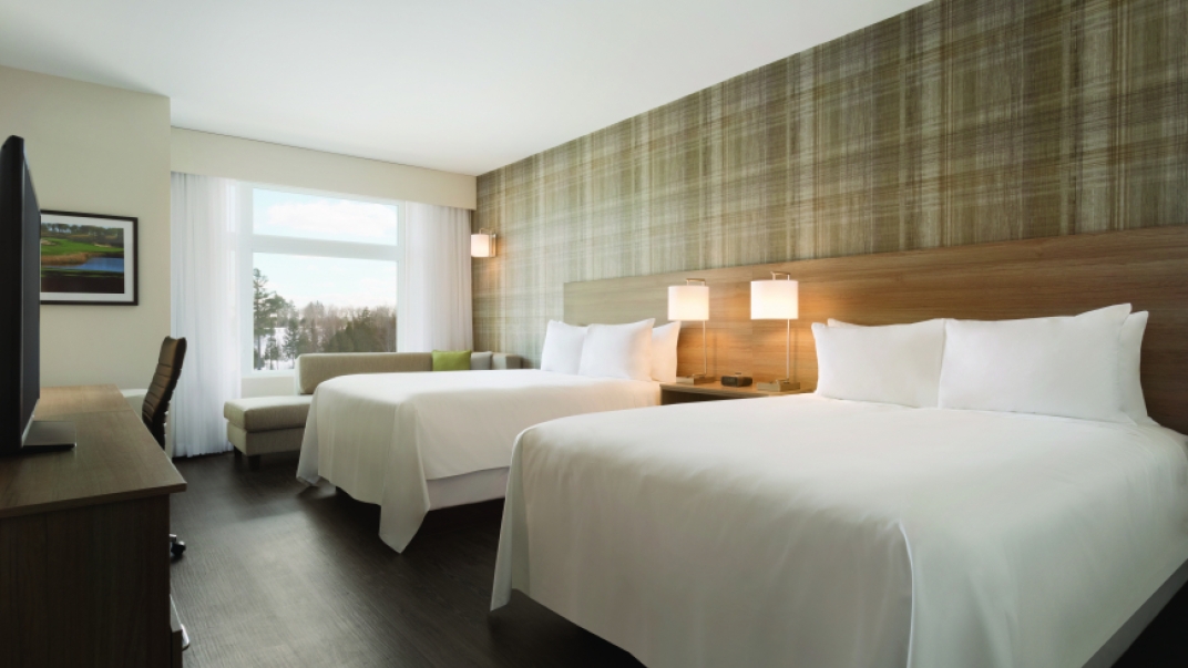 Inside shot of a hotel room with clean white sheets, pillows, and a large window. 