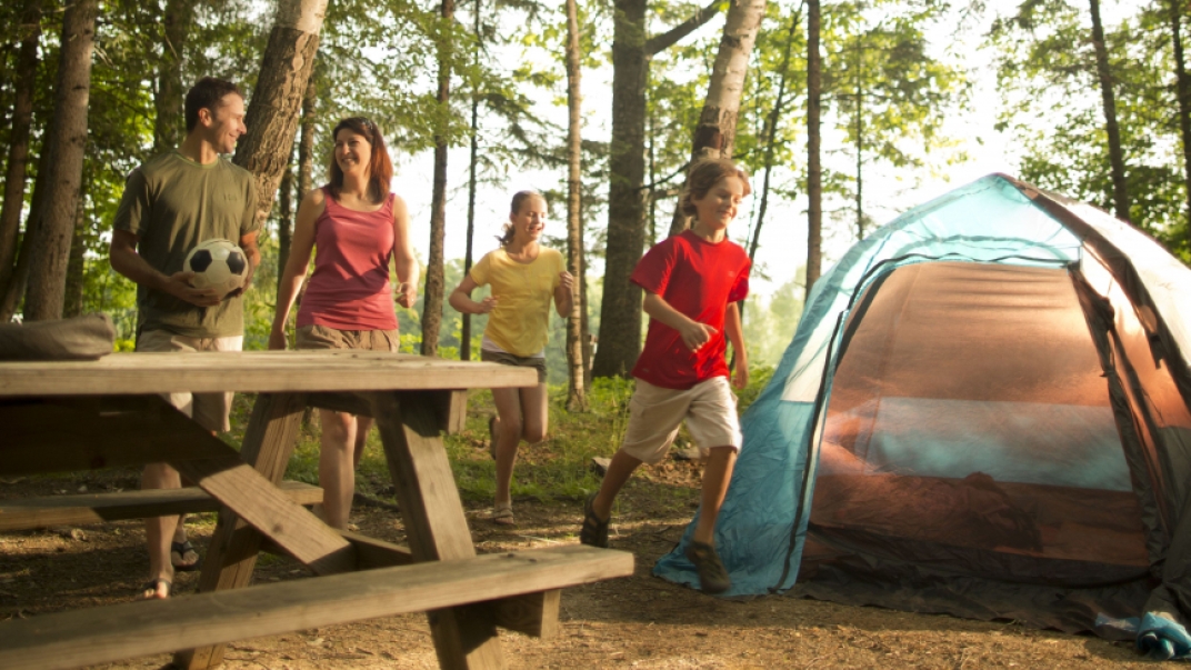 Smiling family playing at a campsite in a forest with a tent and picnic table.