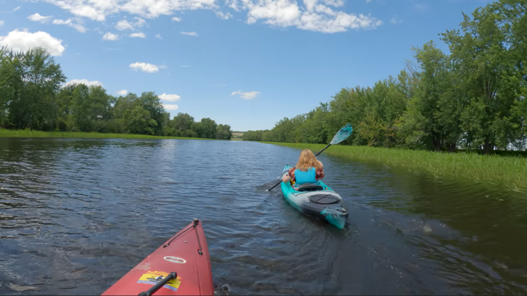 Two kayakers paddle down a narrow blue river towards a bright blue sky with fluffy clouds.