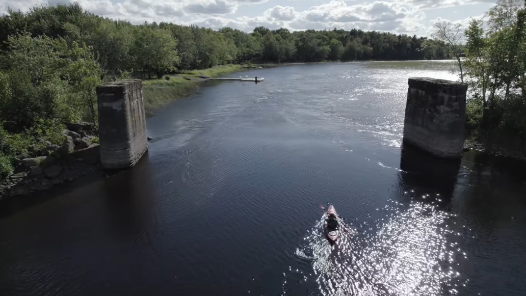 A kayaker paddles their way down a beautiful blue river banked by forests on both sides.