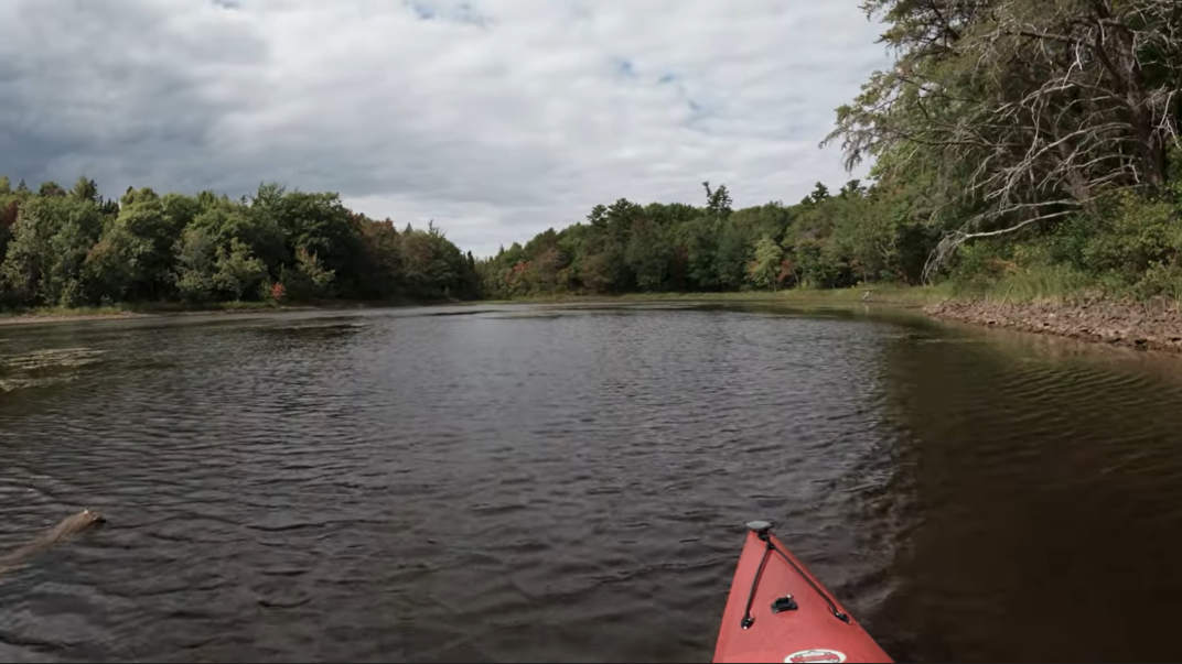 The tip of a red paddle juts out onto a deep blue river banked by sand and trees on a cloudy day