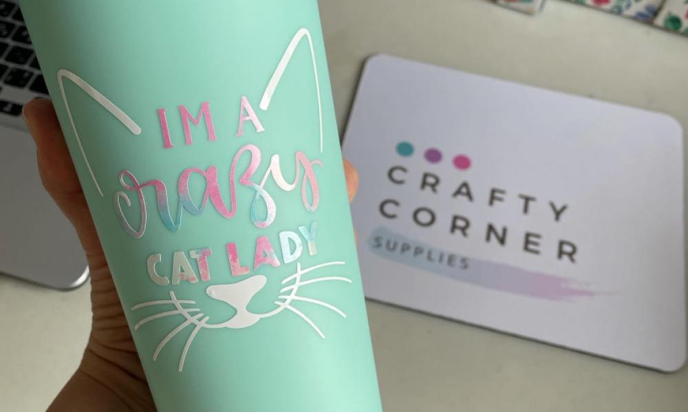 Metal mug with a cat graphic and the words "I am a crazy cat aldy" written on it. In the background there is a mat with the words" Crafty Corner" on it.