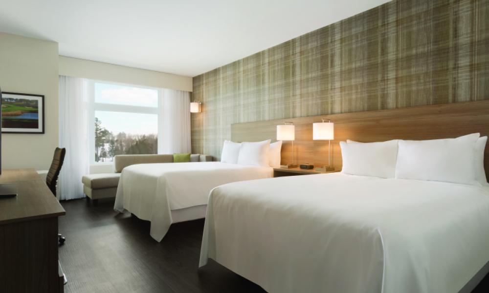 Inside shot of a hotel room with clean white sheets, pillows, and a large window. 