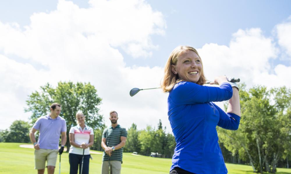 white woman in bright blue long sleeve shirt swings a golf club while her companions watch in the background