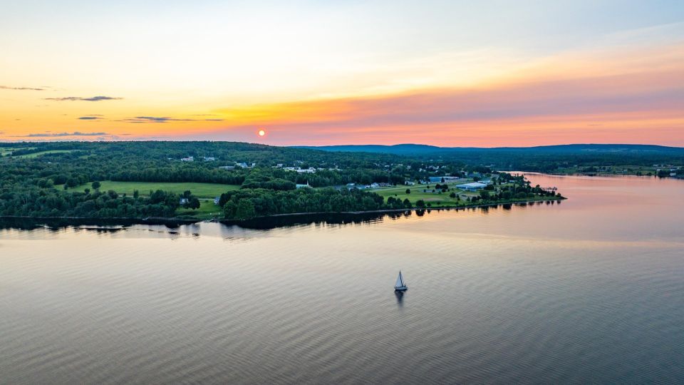 Long shot from above of a sailboat on a river with green forests behind it against an orange sunset.