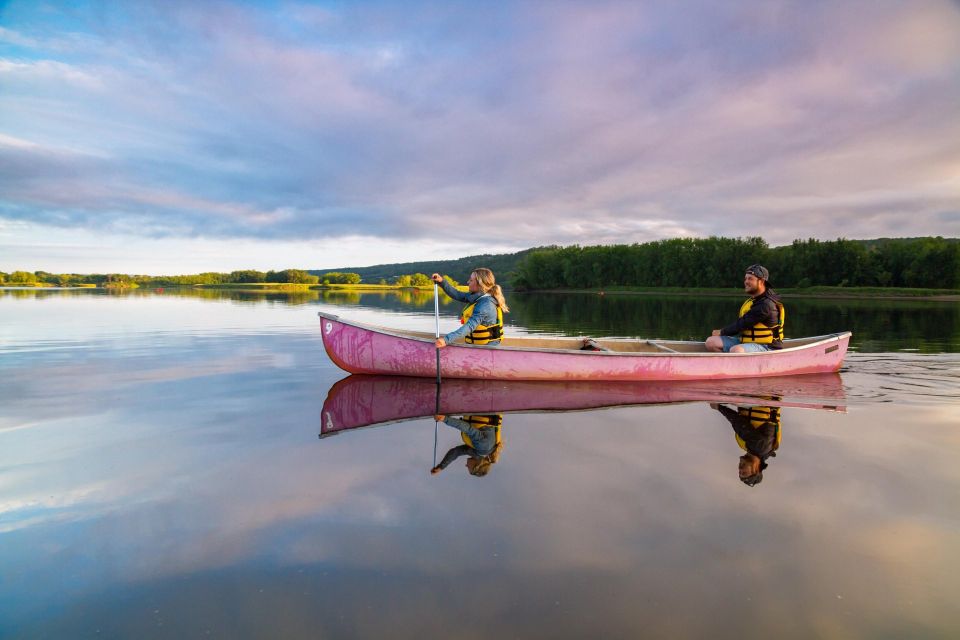 Two people paddle a canoe across calm water. The canoe and sky reflect clearly on the lake’s surface.