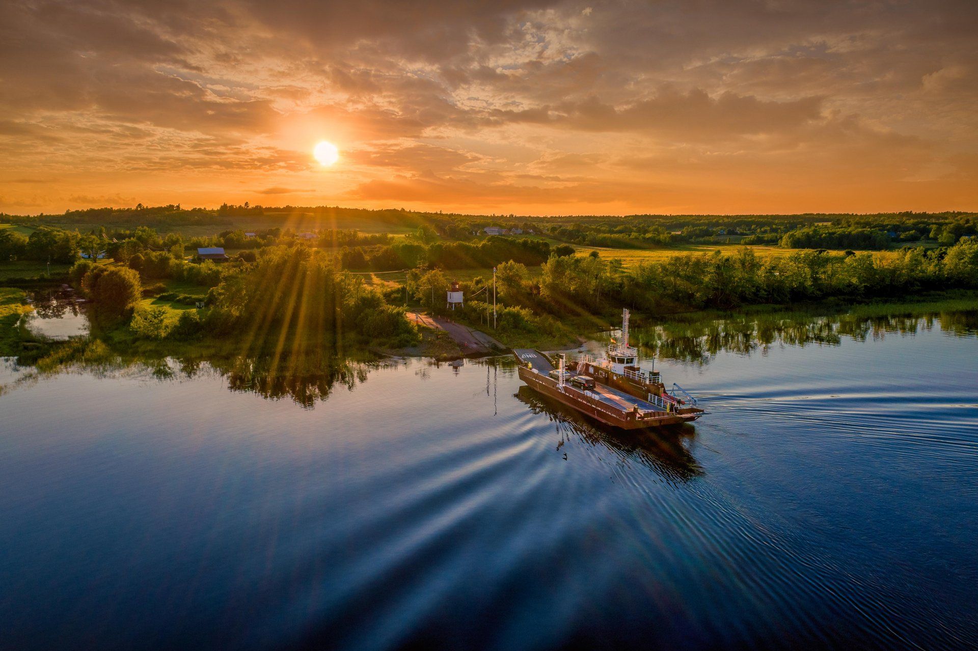 A small ferry crosses a calm river at sunset while golden sunlight streams over a green shoreline and the boat’s wake rippling across the water.