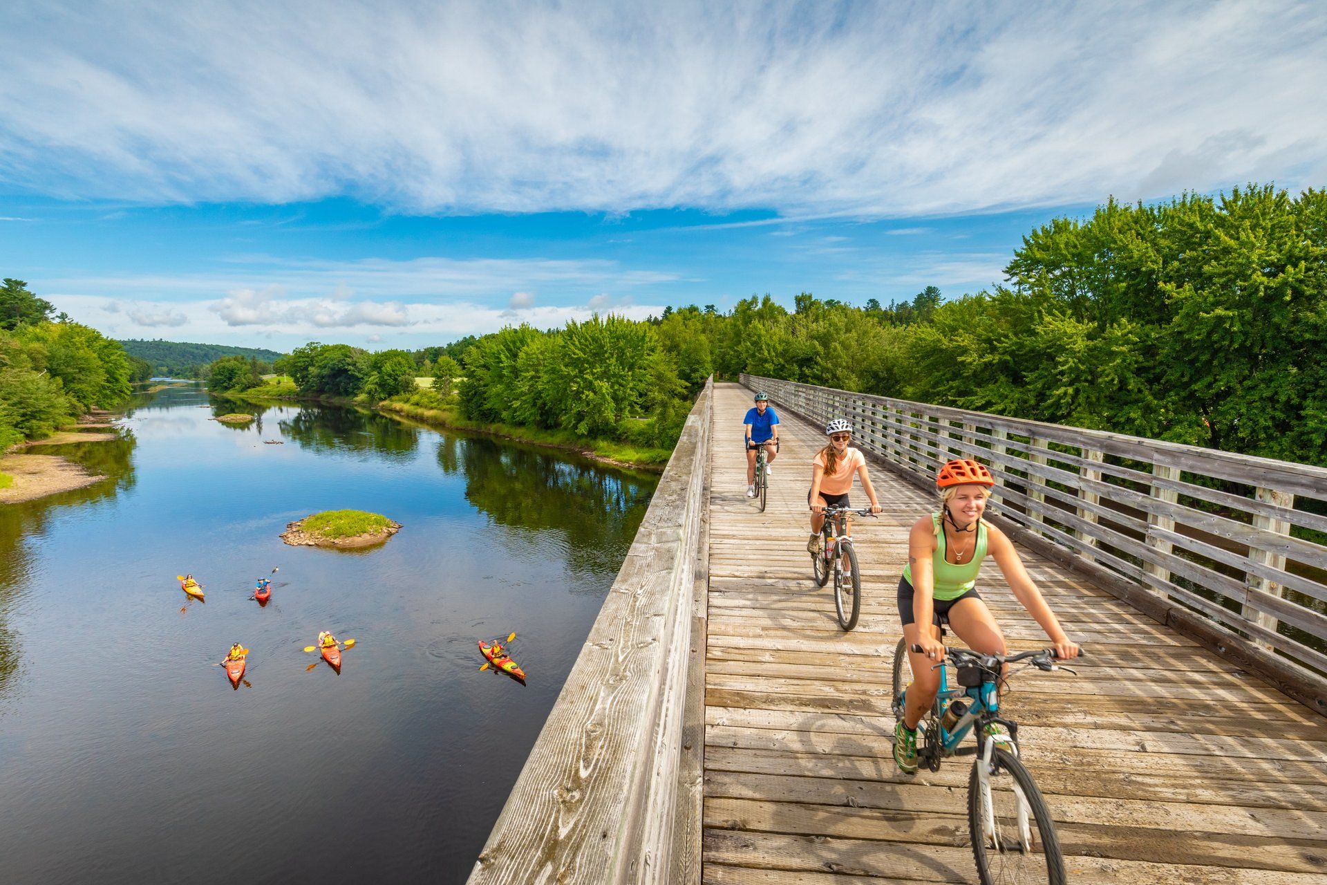 Three cyclists ride across a wooden bridge over a river. Kayakers paddle below on calm water surrounded by green trees.