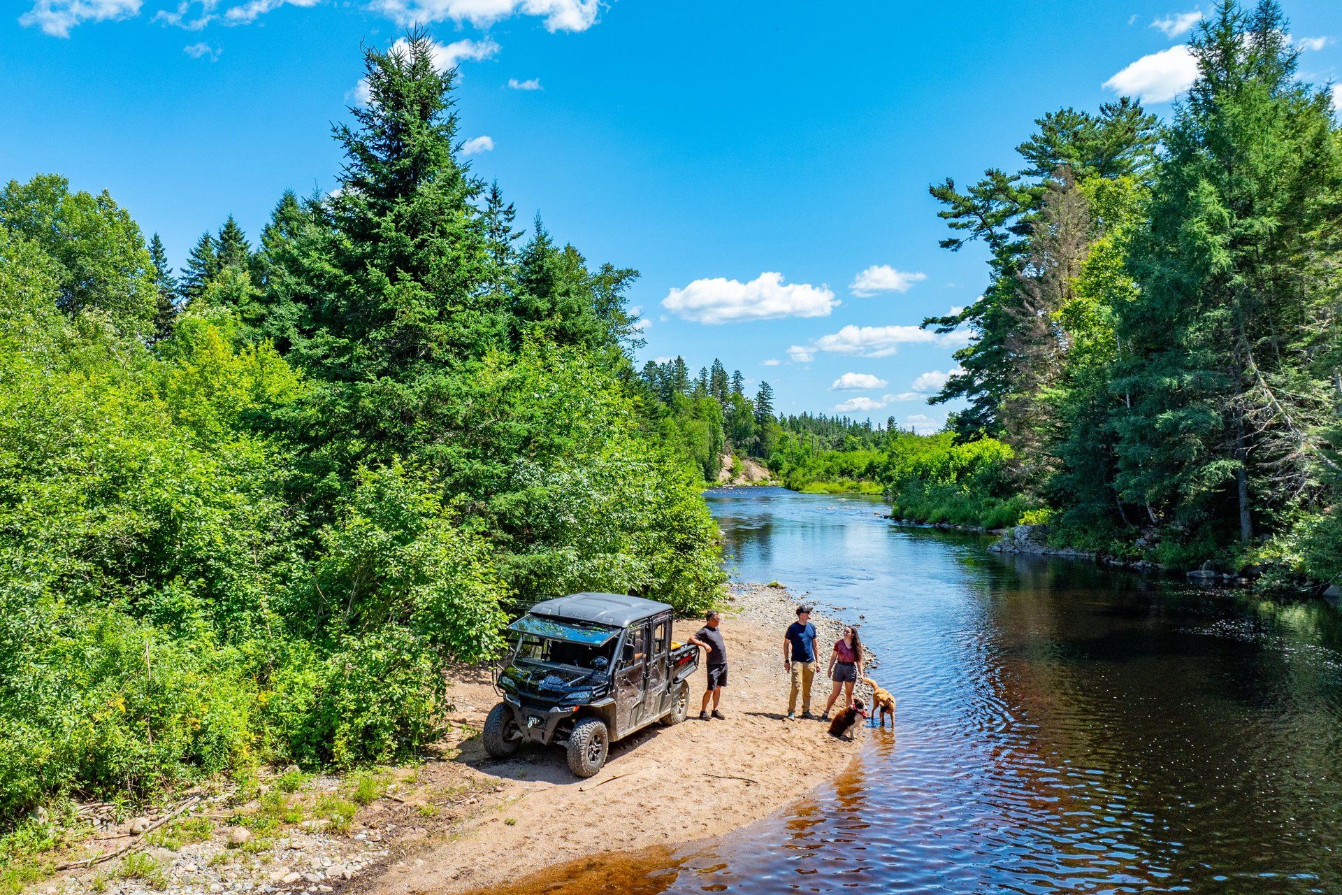 Three people stand beside an ATV on a sandy riverbank. Two dogs play at the water’s edge, surrounded by trees under a blue sky.