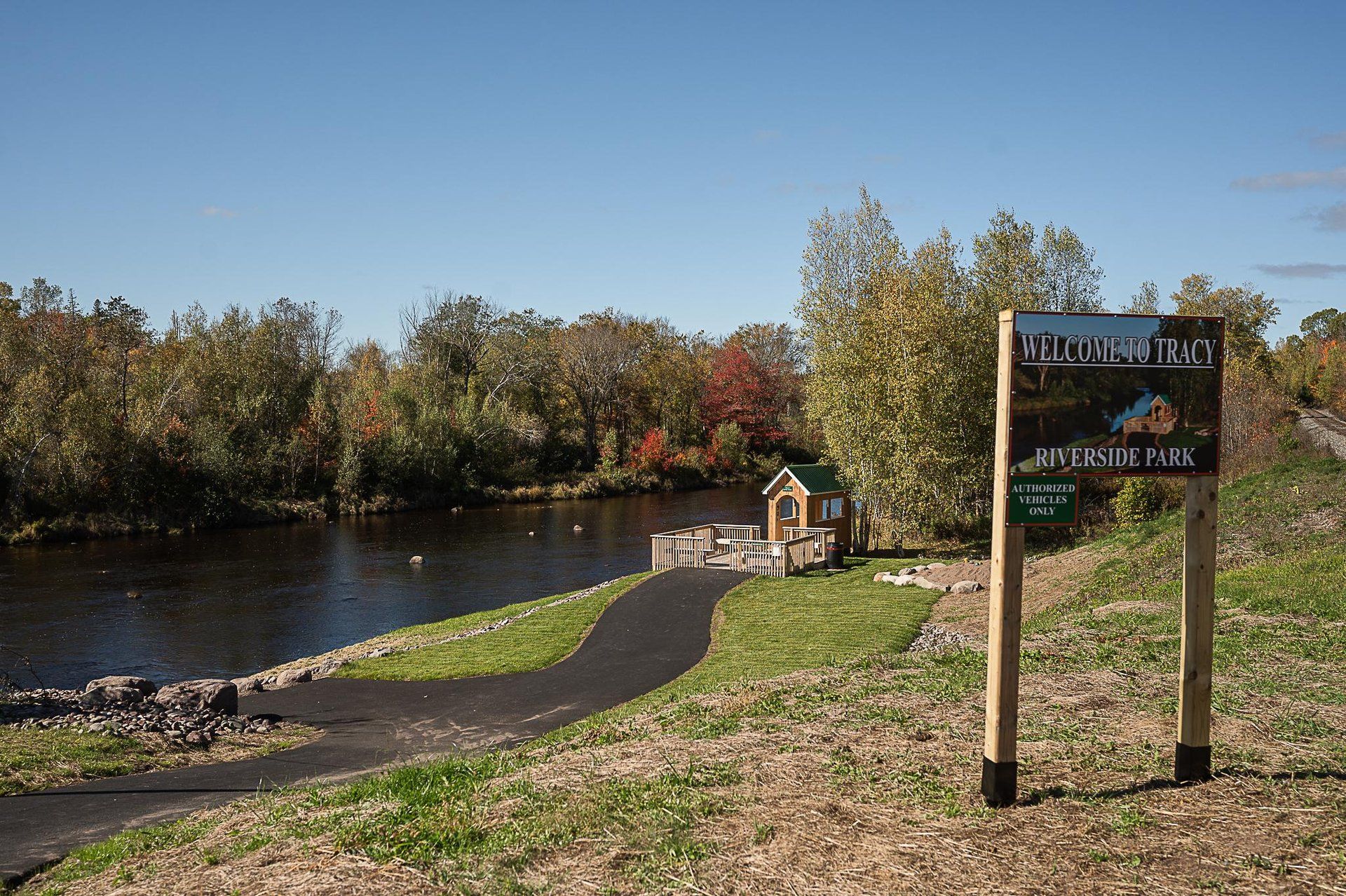 A sign reading “Welcome to Tracy Riverside Park” stands beside a paved path leading to a small deck by a river lined with autumn trees.
